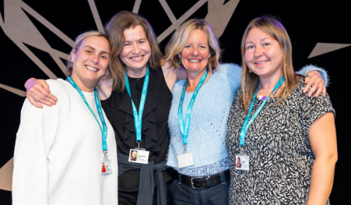Four women stand with their arms around each others shoulders and smile at the camera.